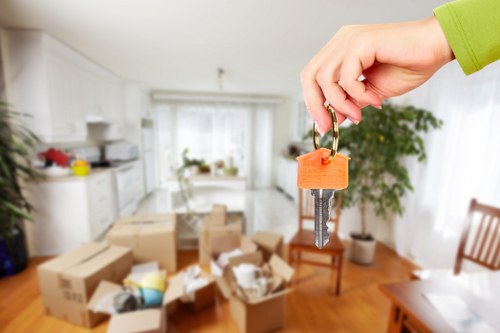 A person's hand, wearing a lime green sleeve, is holding a key attached to an orange plastic keyring, with a background of a bright, well-lit living room featuring white walls, large windows with sheer curtains, a wooden dining table surrounded by chairs, and various cardboard moving boxes scattered across the wooden floor. The boxes are partially open, revealing packing paper, household items, and small appliances packed inside. The room appears to be in a residential house in Edgware, with natural light filling the space, and some green potted plants visible near the windows, hinting at a house prepared for a relocation or move managed by Edgware Man with Van.