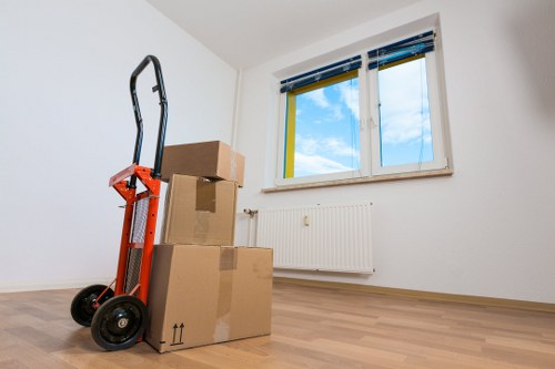 In a well-lit, empty room with white walls and light wooden flooring, three large cardboard boxes of varying sizes are stacked near a radiator beneath a window with blue and yellow blinds. The boxes are closed and sealed, showing typical brown corrugated cardboard with black printed symbols indicating fragile contents and upright positioning. A red hand truck or dolly with black handles and wheels is positioned upright beside the boxes, suggesting recent use for moving or loading. Outside the window, a clear blue sky with some clouds is visible, indicating daytime. The scene appears to be part of a house relocation or removal process, with the absence of furniture and minimal decor emphasizing that the space is either prepared for moving activities or recently vacated. The presence of the boxes and hand truck references the services offered by Edgware Man with Van, highlighting their local house removals expertise in the Edgware area, contained within the vicinity of NW London.