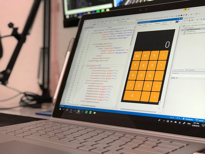 A person with short dark hair, dressed in a light blue shirt, is seated at a desk or table reading an open laptop displaying lines of color-coded programming code, likely related to IT or software dev