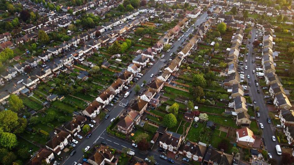 An aerial view of a suburban residential neighborhood showing rows of detached and semi-detached houses with gardens, driveways, and tree-lined streets. The image captures several parked cars along th