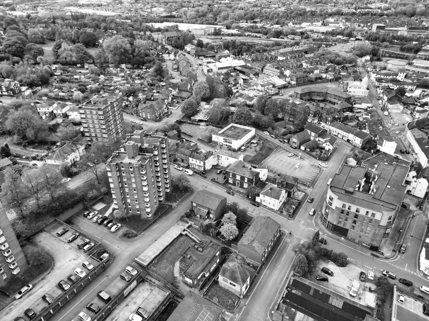 An aerial view of a suburban residential neighborhood showing rows of detached and semi-detached houses with gardens, driveways, and tree-lined streets. The image captures several parked cars along th